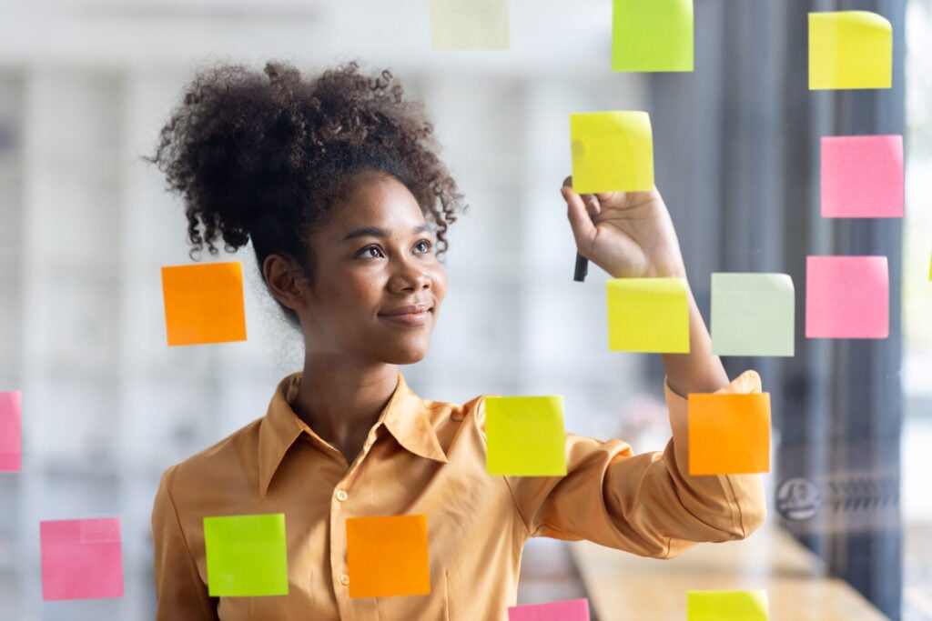 Une jeune femme souriante écrit sur des post-its colorés collés sur une vitre, en pleine réflexion créative.