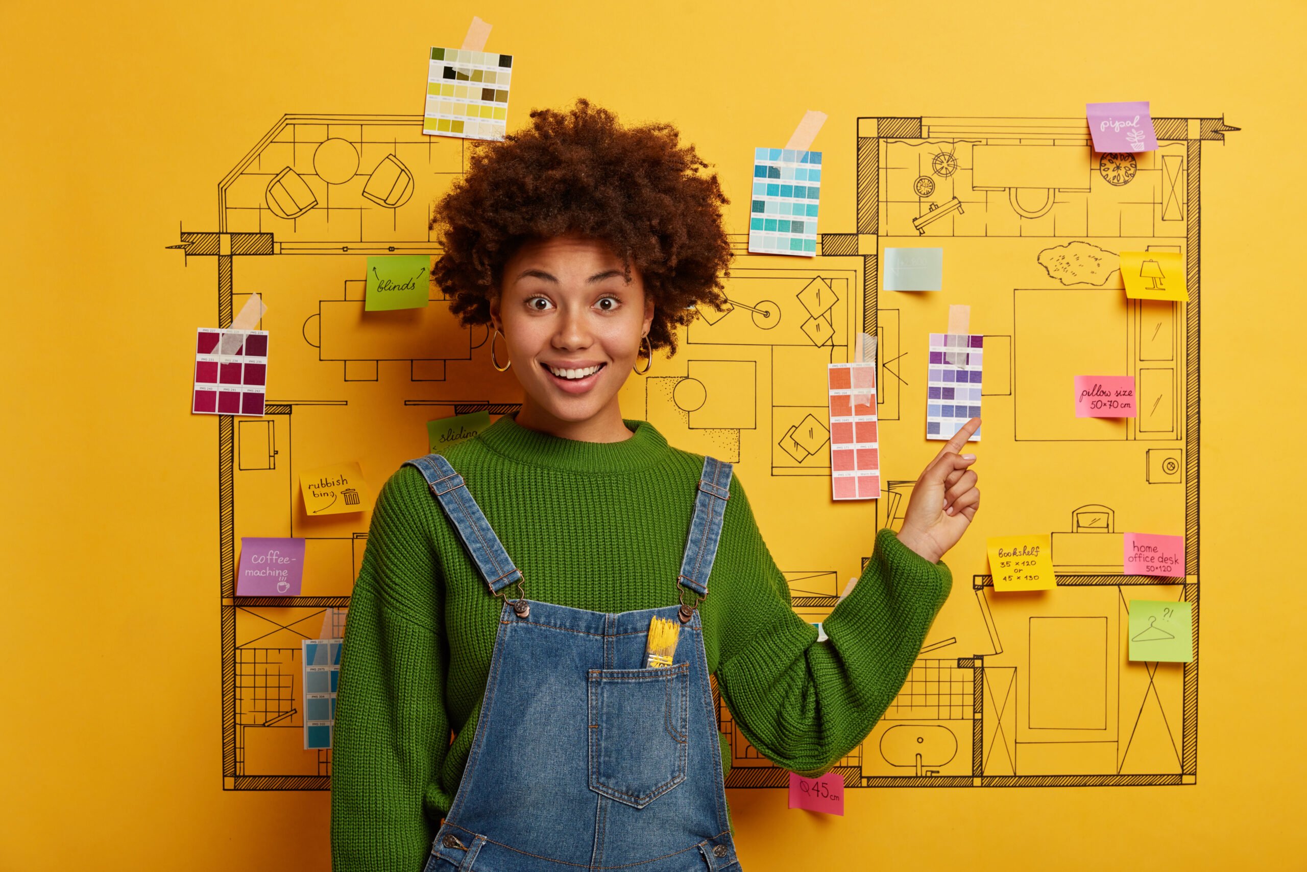 Une femme souriante en salopette, debout devant un plan d’aménagement affiché sur un mur jaune, entourée de nuanciers de couleurs et de post-its.