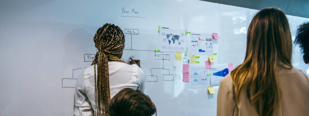 Personne dessinant un schéma sur un grand tableau blanc avec des post-its colorés et des documents affichés.