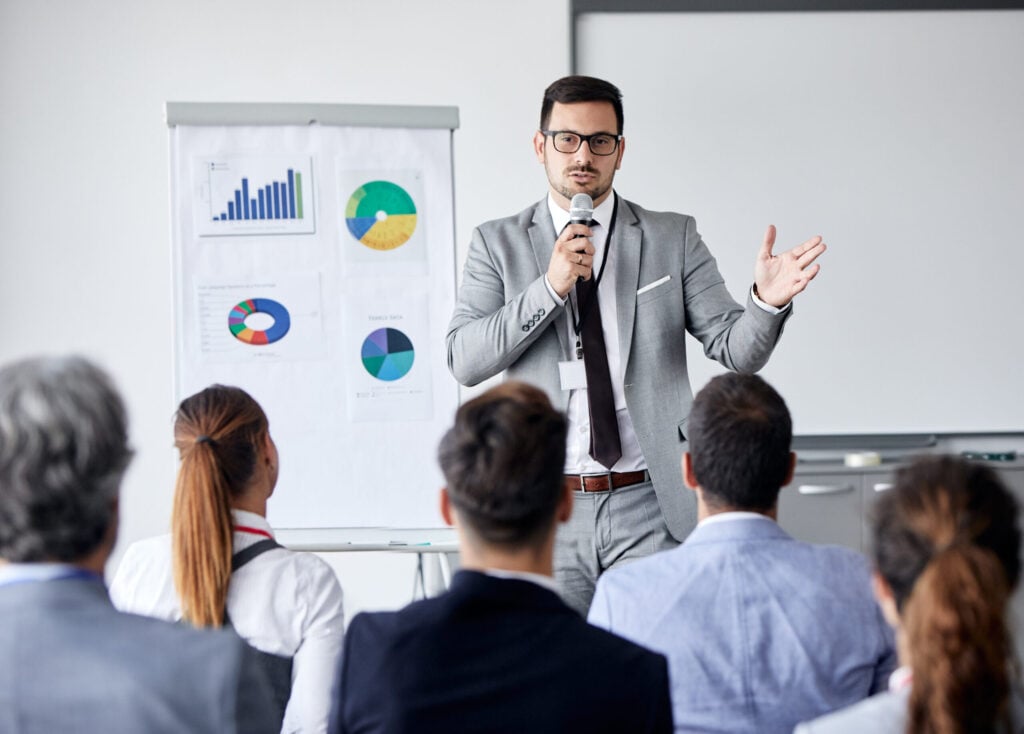 Manager en costume s’adressant à un groupe lors d’une présentation avec un micro, devant un paperboard.